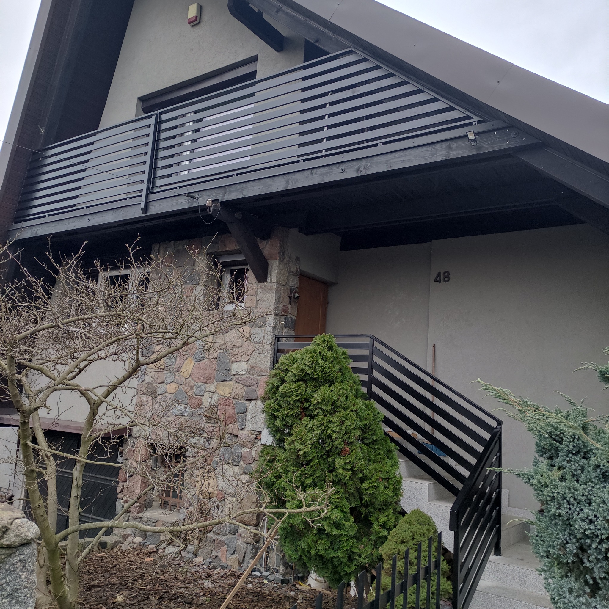 Dom z balkonem i schodami, zabezpieczonymi czarnymi, poziomymi balustradami. Kamienna elewacja do połowy wysokości. Numer domu '48' widoczny na ścianie.