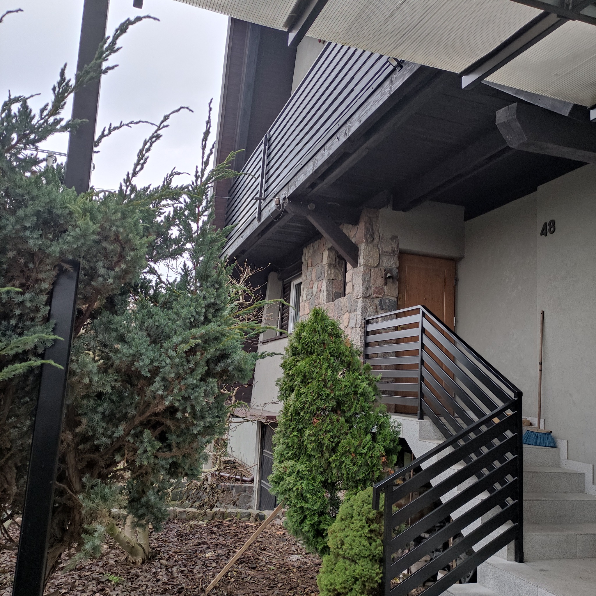 Nowoczesna balustrada schodowa z czarnych, poziomych profili, prowadząca do wejścia domu z kamienną elewacją i numerem 48. Widok z poziomu ogrodu, częściowo zasłonięta przez roślinność.