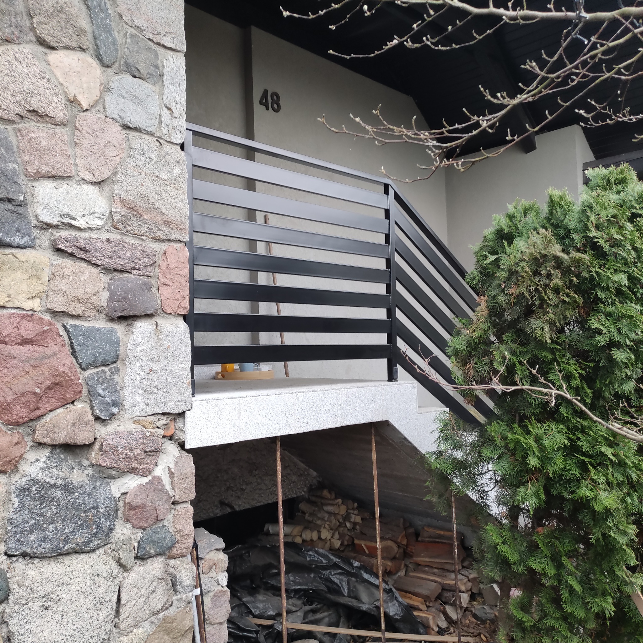Nowoczesna, pozioma balustrada schodowa w kolorze antracytowym, zamontowana przy wejściu do domu z elewacją z kamienia. Widoczny numer domu '48'.