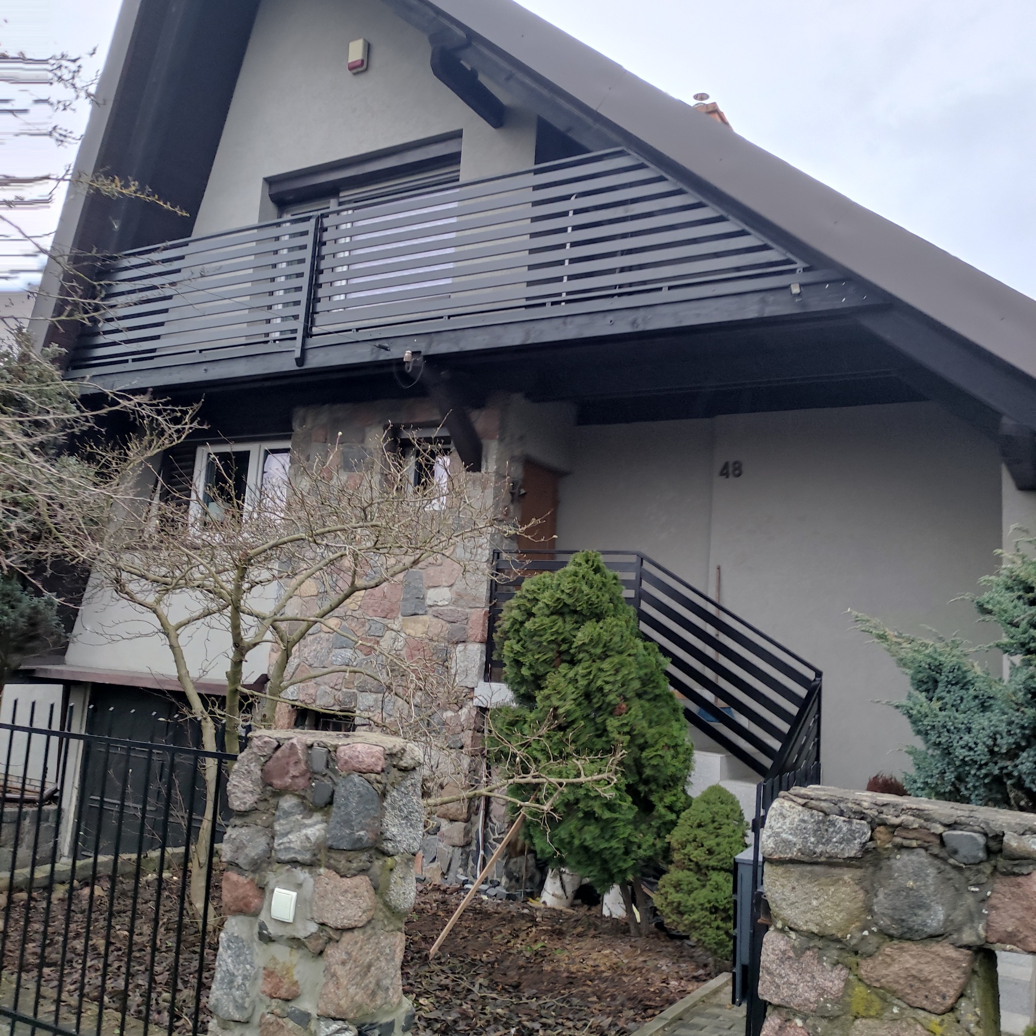 Dom z balkonem i schodami z czarnymi, poziomymi balustradami. Kamienne wykończenie elewacji i ogrodzenia. Numer domu '48' widoczny na ścianie.