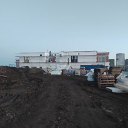 Usługi budowlane Grzegorz Deda - Budynek w trakcie prac wykończeniowych, rusztowania, materiały budowlane, ziemia. Widoczne osoby pracujące na wysokościach. Niebo bezchmurne.