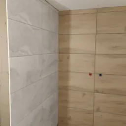 Wykończona ściana z płytek imitujących drewno i marmur, widoczne podejścia wodne. Jasne kolory, minimalistyczny design.