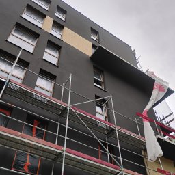 Usługi budowlane Grzegorz Deda - Elewacja budynku w trakcie prac wykończeniowych, widoczne rusztowania i fragment ocieplenia. Okna zabezpieczone folią, pracownik na górze budynku.