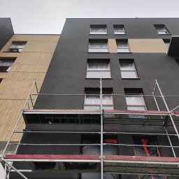 Usługi budowlane Grzegorz Deda - Elewacja budynku w trakcie malowania na szaro i beżowo, okna zabezpieczone folią, widoczne rusztowania. Ujęcie z dołu.
