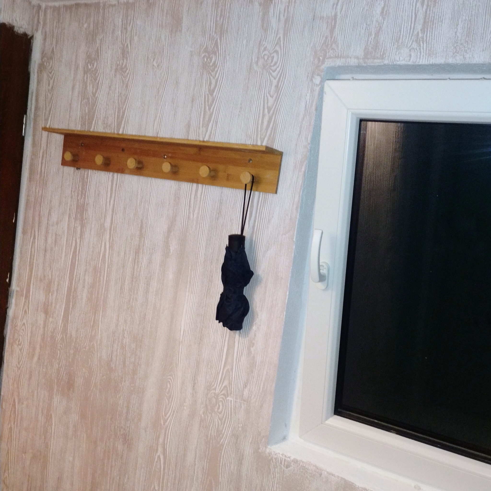 Ściana z tapetą imitującą drewno, drewniany wieszak z parasolem i białe okno. Widoczne niedoskonałości wykończenia przy oknie.