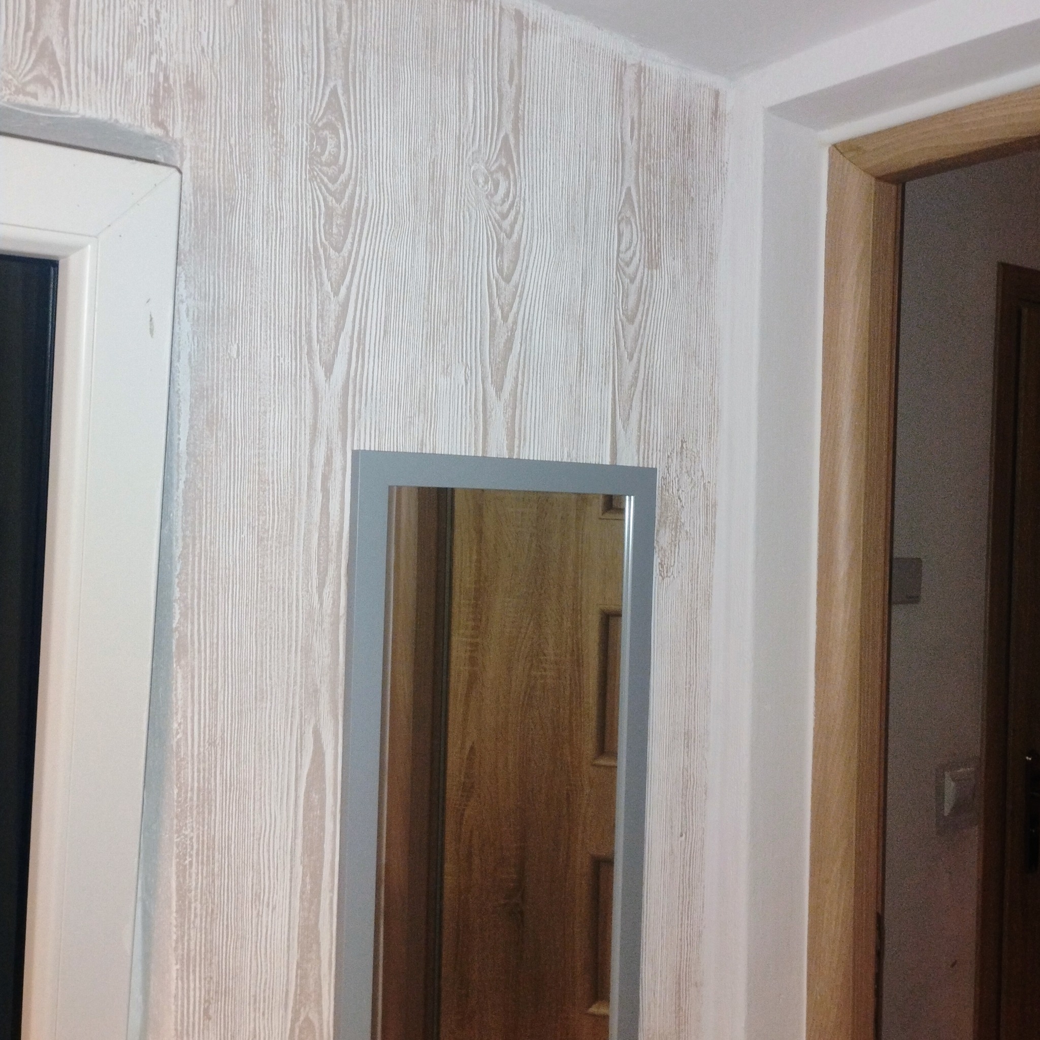 Ściana z tapetą imitującą drewno, fragment futryny i lustro w szarej ramie. Widok na wewnętrzne drzwi z korytarza. Realizacja z detalami wykończenia w jasnej tonacji.