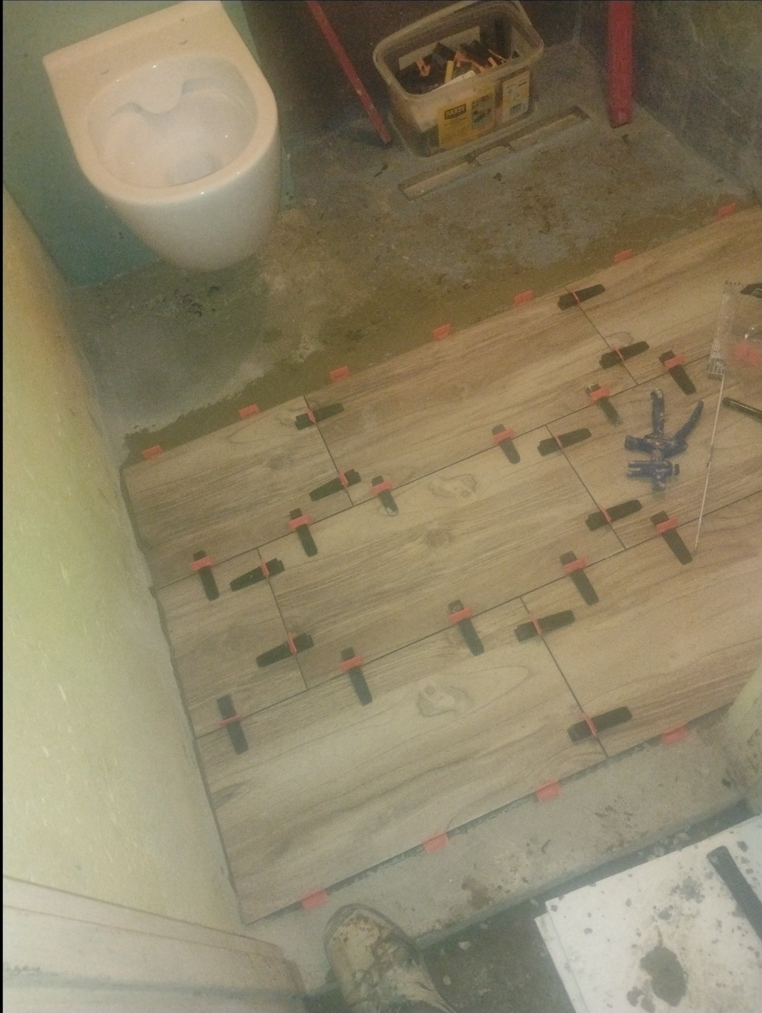 Układanie płytek imitujących drewno w łazience, z widocznymi elementami poziomującymi. W tle toaleta i narzędzia glazurnicze.