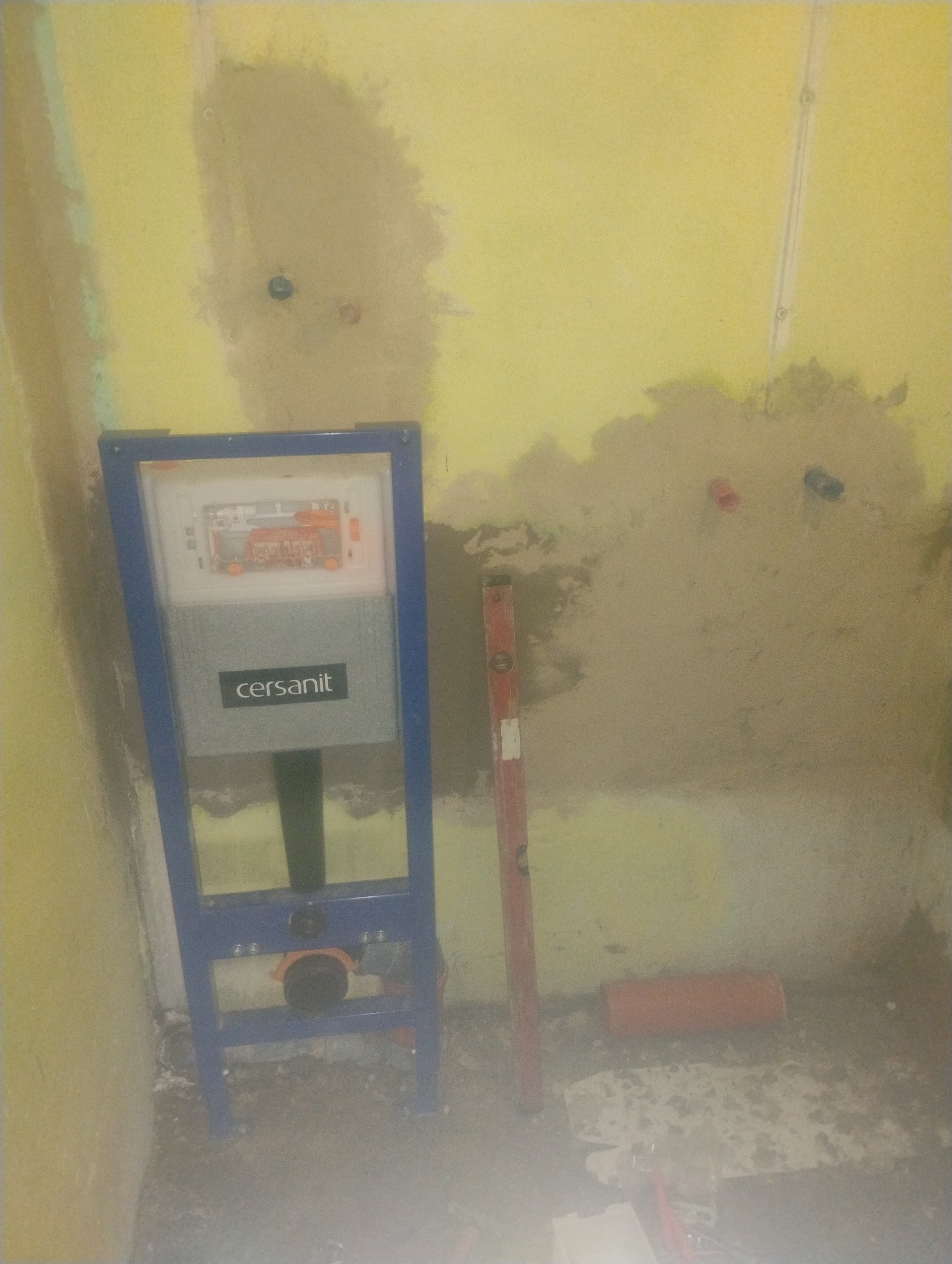 Stelaż podtynkowy Cersanit zamontowany przy żółtej ścianie w trakcie remontu łazienki. Widoczny poziomica i fragment rury kanalizacyjnej.