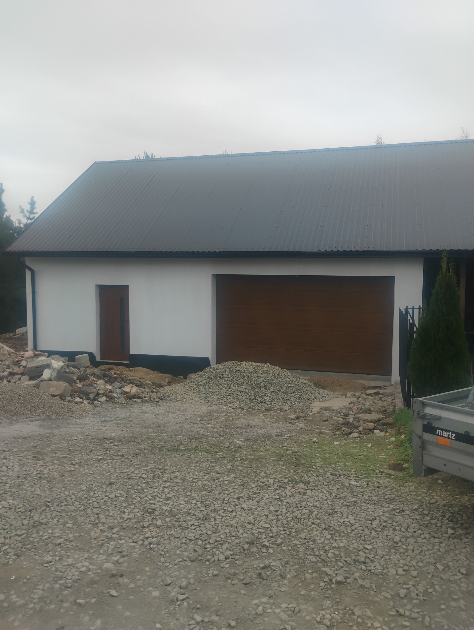 Nowoczesny garaż z brązową bramą segmentową i białymi ścianami, na tle szarego nieba. Wokół rozsypany gruz i kamienie.