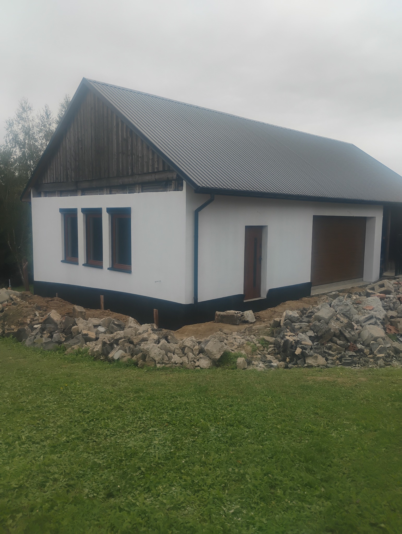 Biały budynek z szarym dachem, brązowymi oknami i drzwiami garażowymi w trakcie wykończenia, z gruzem wokół fundamentów, na tle zielonej trawy i pochmurnego nieba.