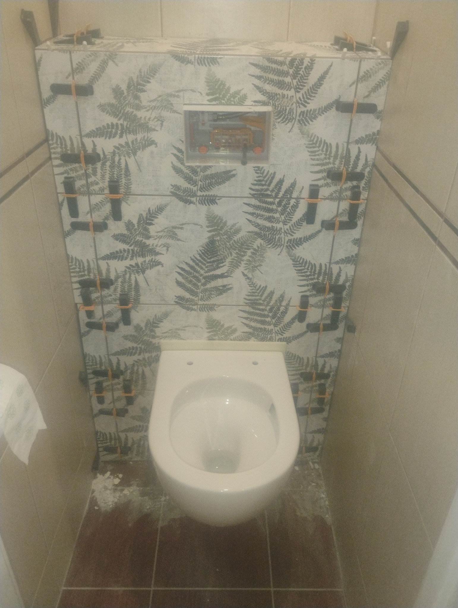 Montaż płytek z motywem paproci wokół toalety podtynkowej, z użyciem krzyżyków dystansowych. Widoczne ślady zaprawy na podłodze.