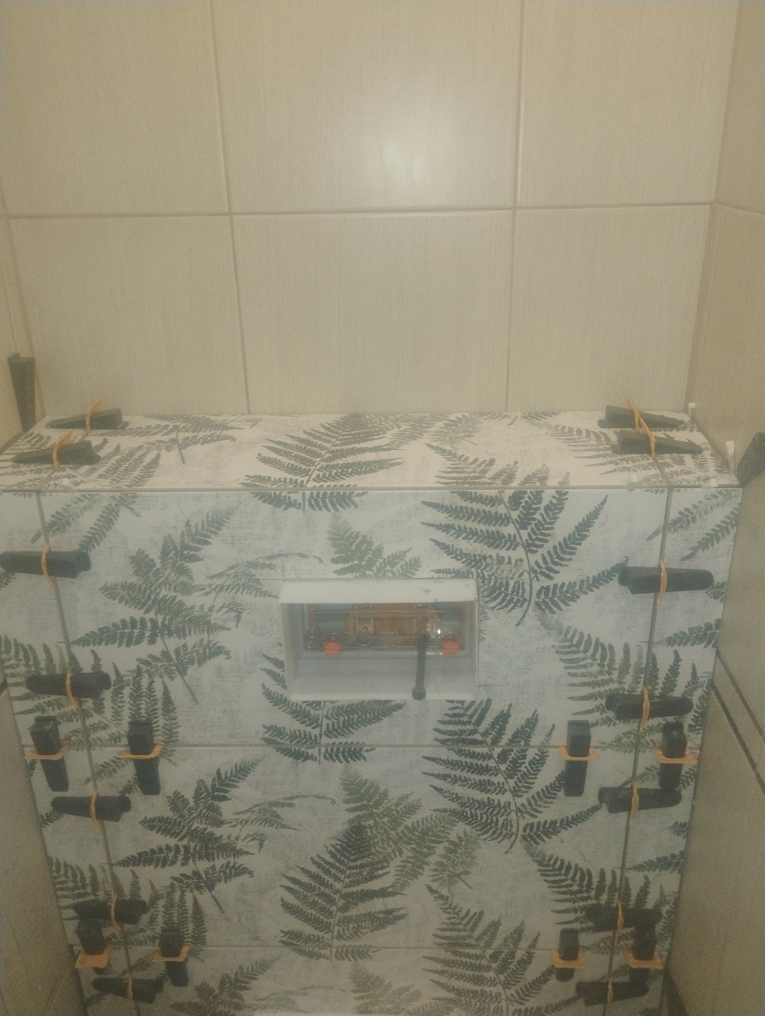 Obudowa stelaża WC z płytek w roślinny wzór, z użyciem klipsów poziomujących. Widoczne jasne płytki na ścianie.