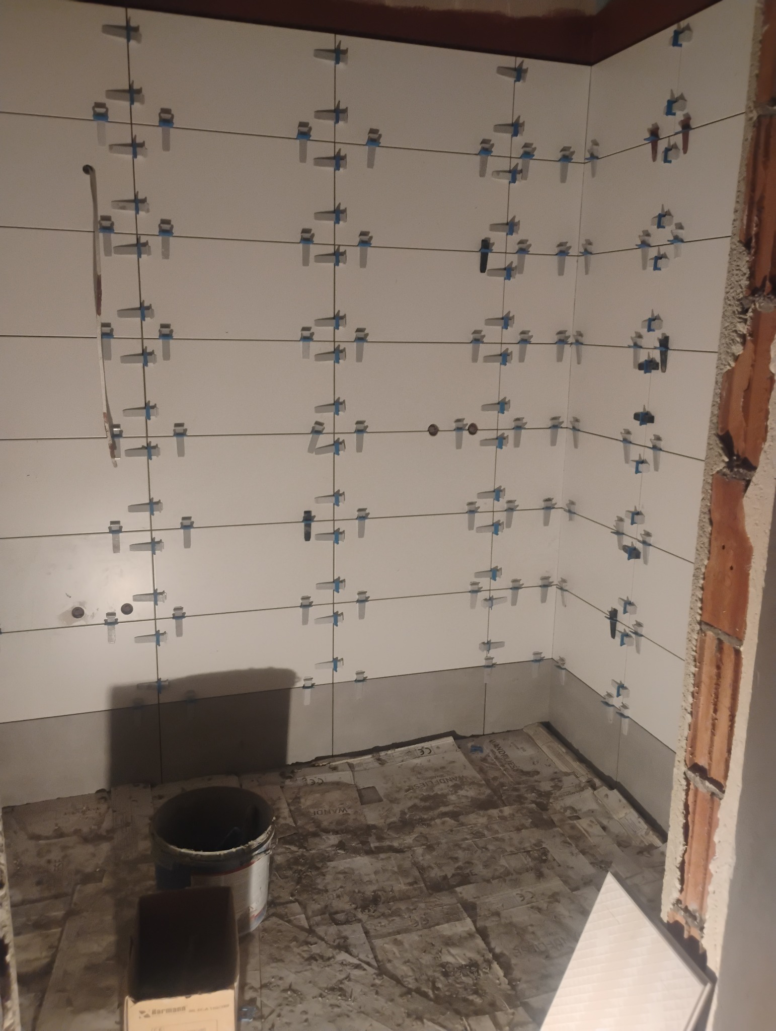 Ściany łazienki w trakcie układania białych płytek, widoczne krzyżyki dystansowe, surowy mur w rogu. Na podłodze kartony ochronne i wiadro.