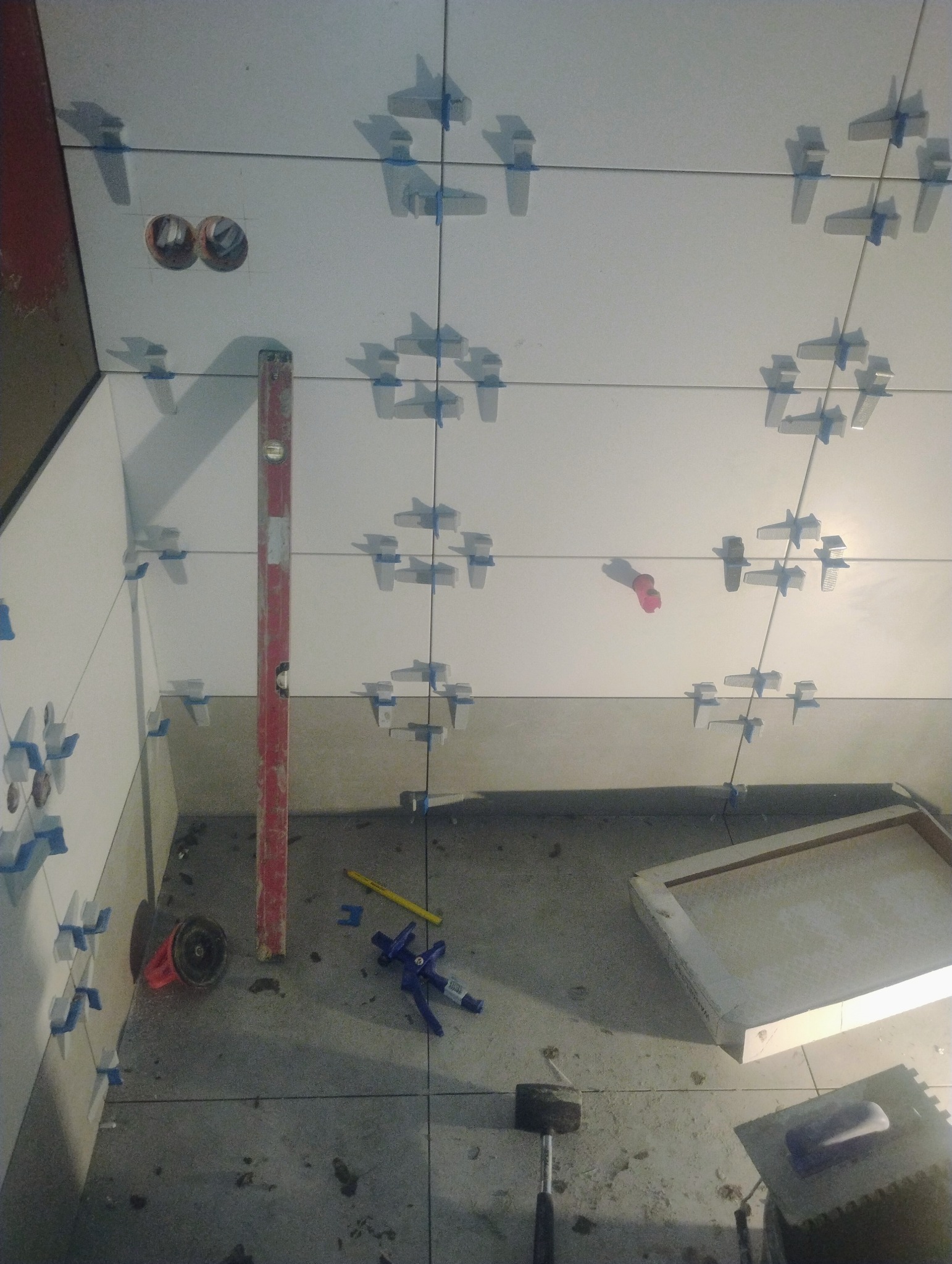 Białe płytki ścienne z niebieskimi krzyżykami dystansowymi, poziomica i narzędzia glazurnicze na podłodze. Widoczne otwory na instalacje.