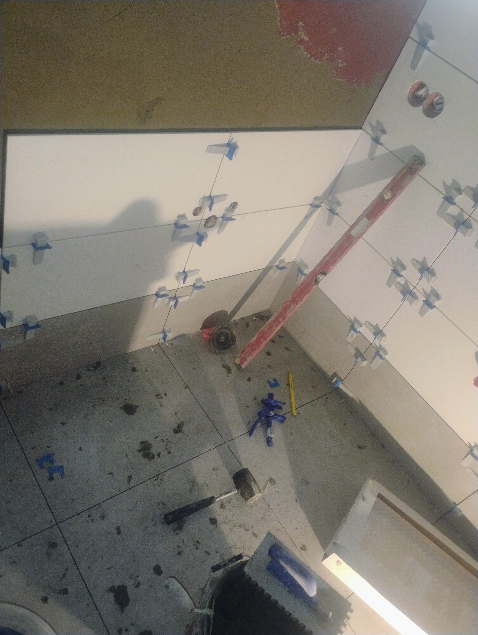 Układanie białych płytek w rogu pomieszczenia, widoczne krzyżyki dystansowe, poziomica i narzędzia glazurnicze. Prace w toku, surowy stan podłoża.