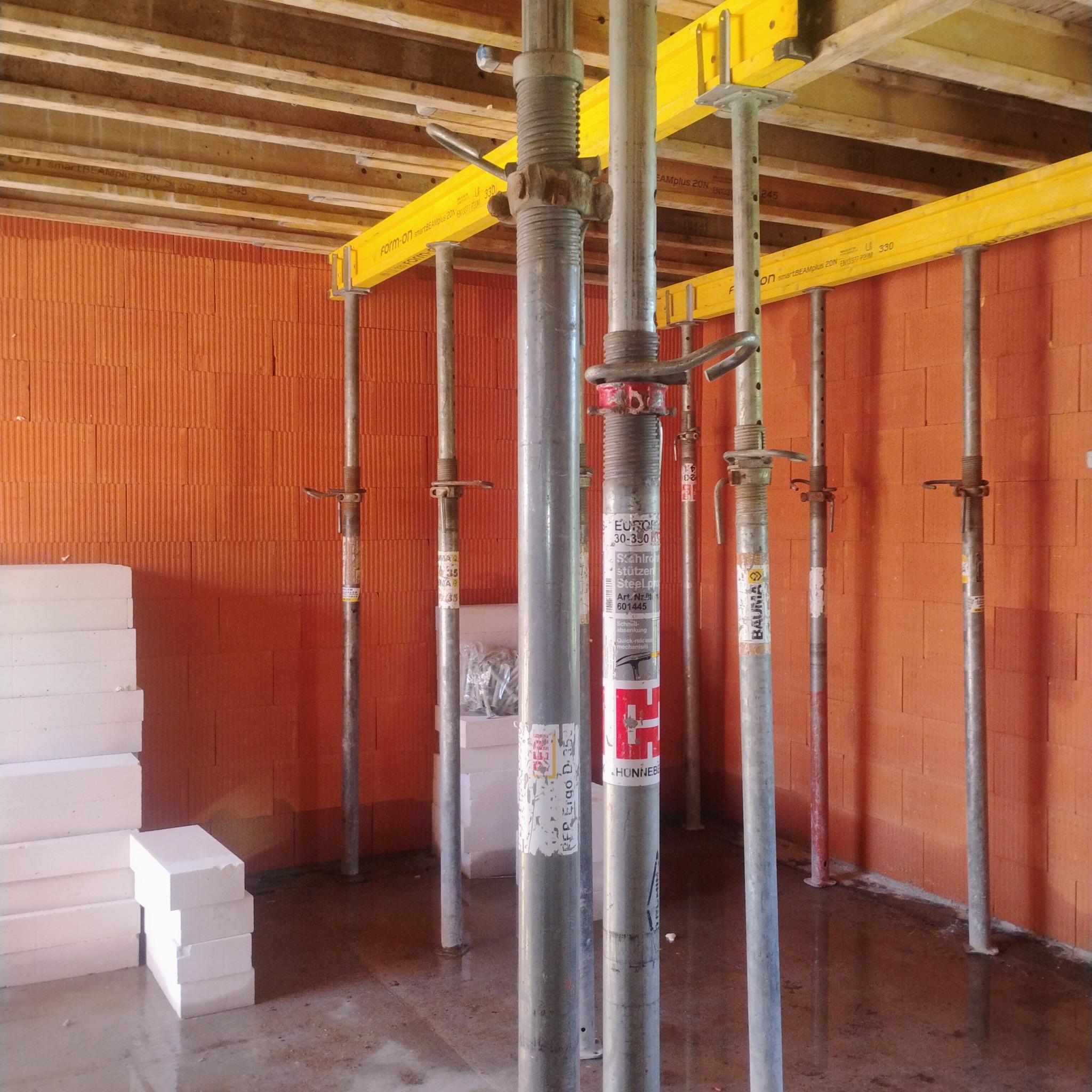 Wnętrze w trakcie budowy: stalowe podpory z żółtymi belkami podtrzymują drewniany strop, widoczne bloczki z betonu komórkowego i ceglana ściana. Mokra posadzka.