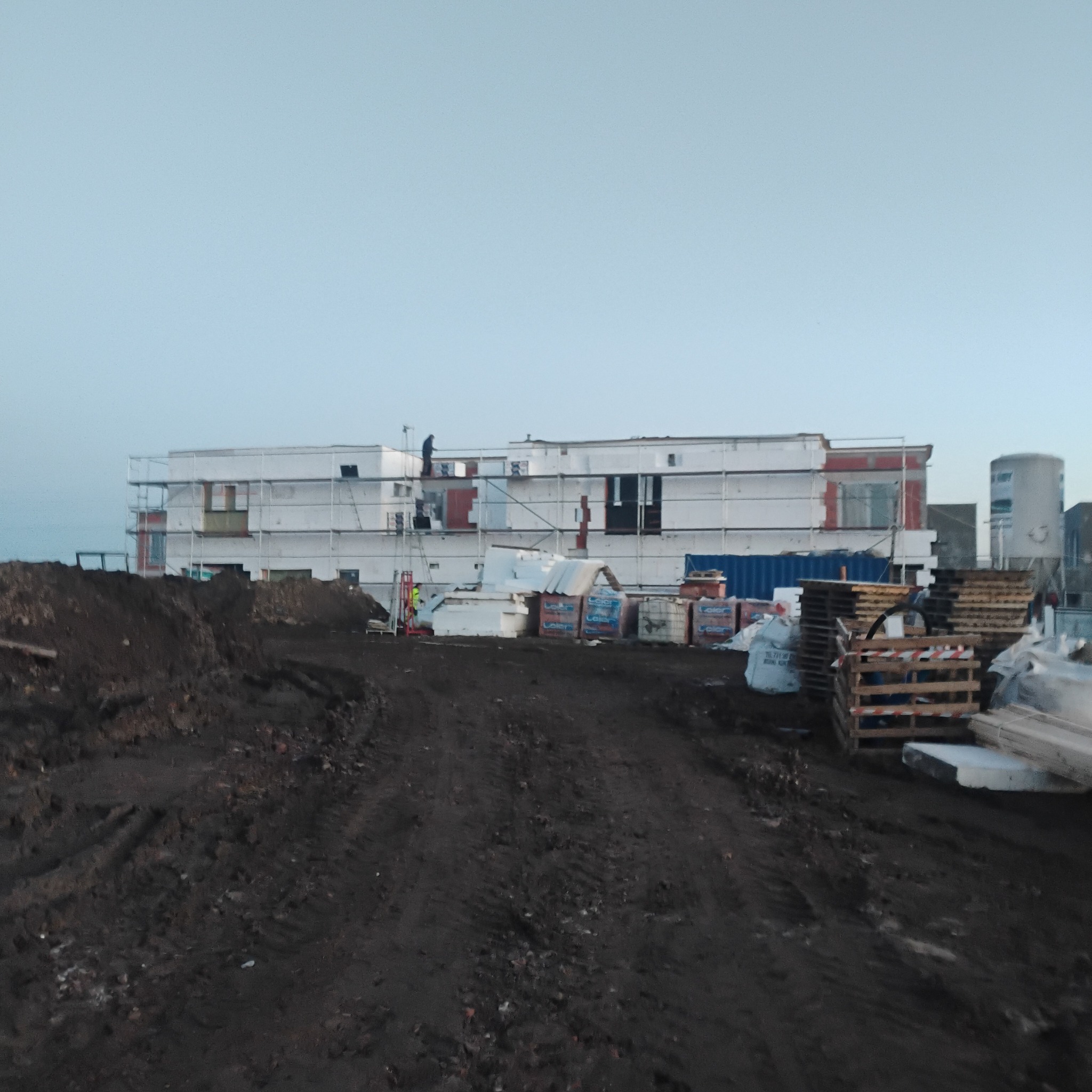 Budynek w trakcie prac wykończeniowych, rusztowania, materiały budowlane, ziemia. Widoczne osoby pracujące na wysokościach. Niebo bezchmurne.