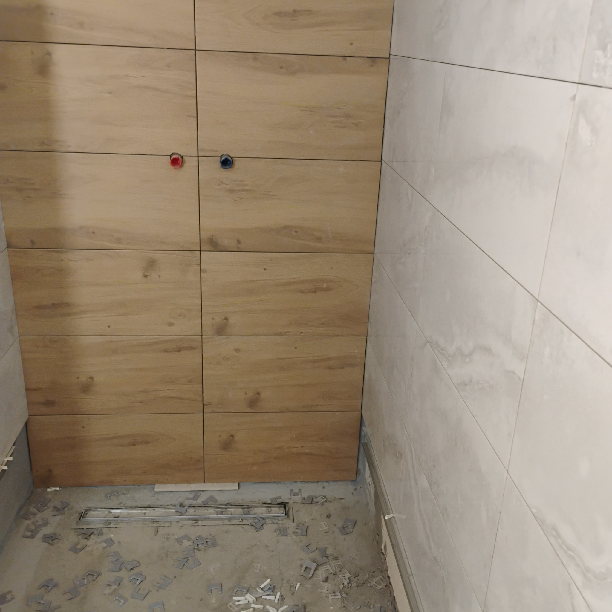 Ściana wyłożona płytkami imitującymi drewno, z widocznymi otworami na armaturę. Na podłodze odpływ liniowy i elementy montażowe. Częściowo wykończona ściana z jasnych płytek.