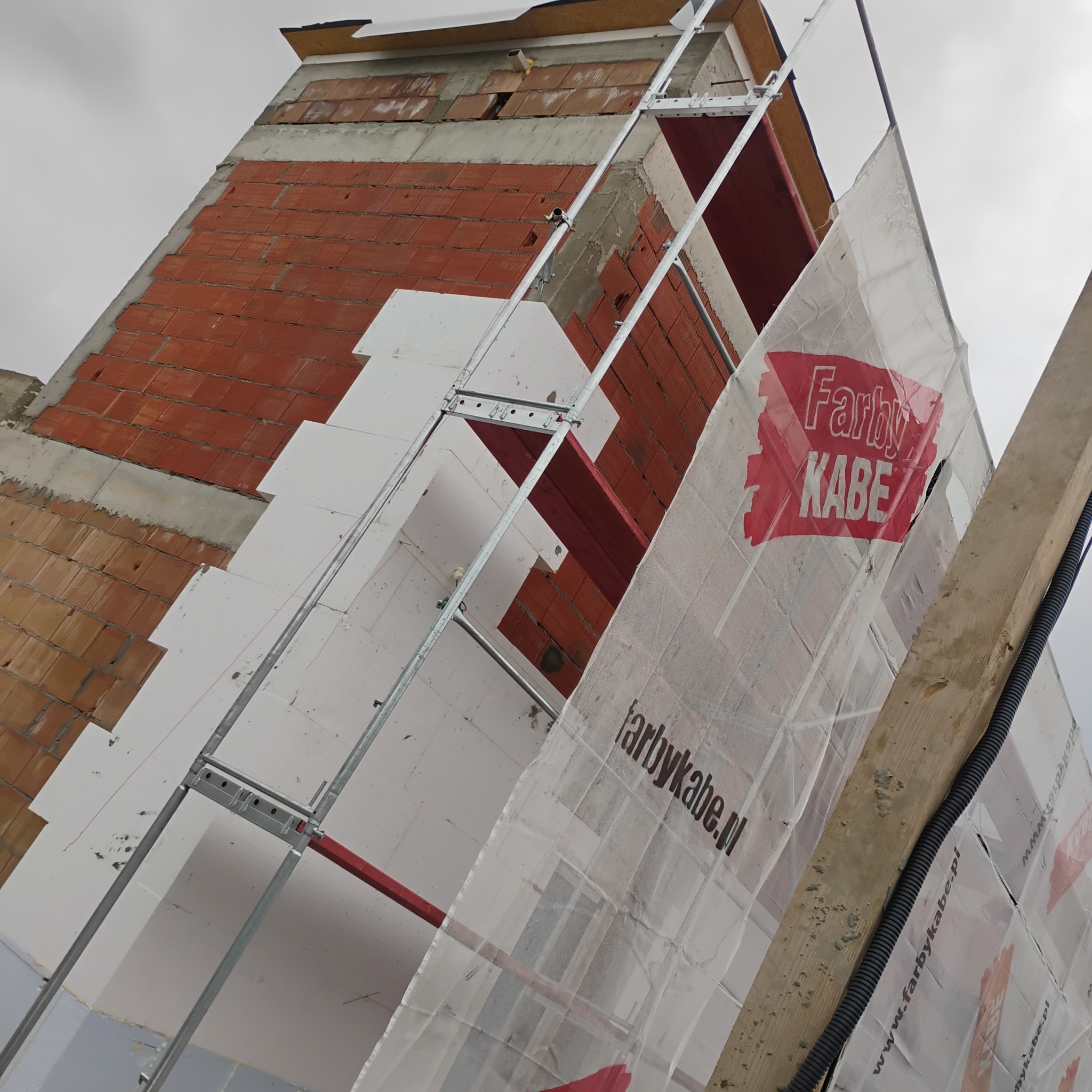 Ocieplenie budynku z rusztowaniem i siatką ochronną z logo Farby Kabe, widok od dołu, na tle pochmurnego nieba. Fragment ściany z czerwonej cegły.