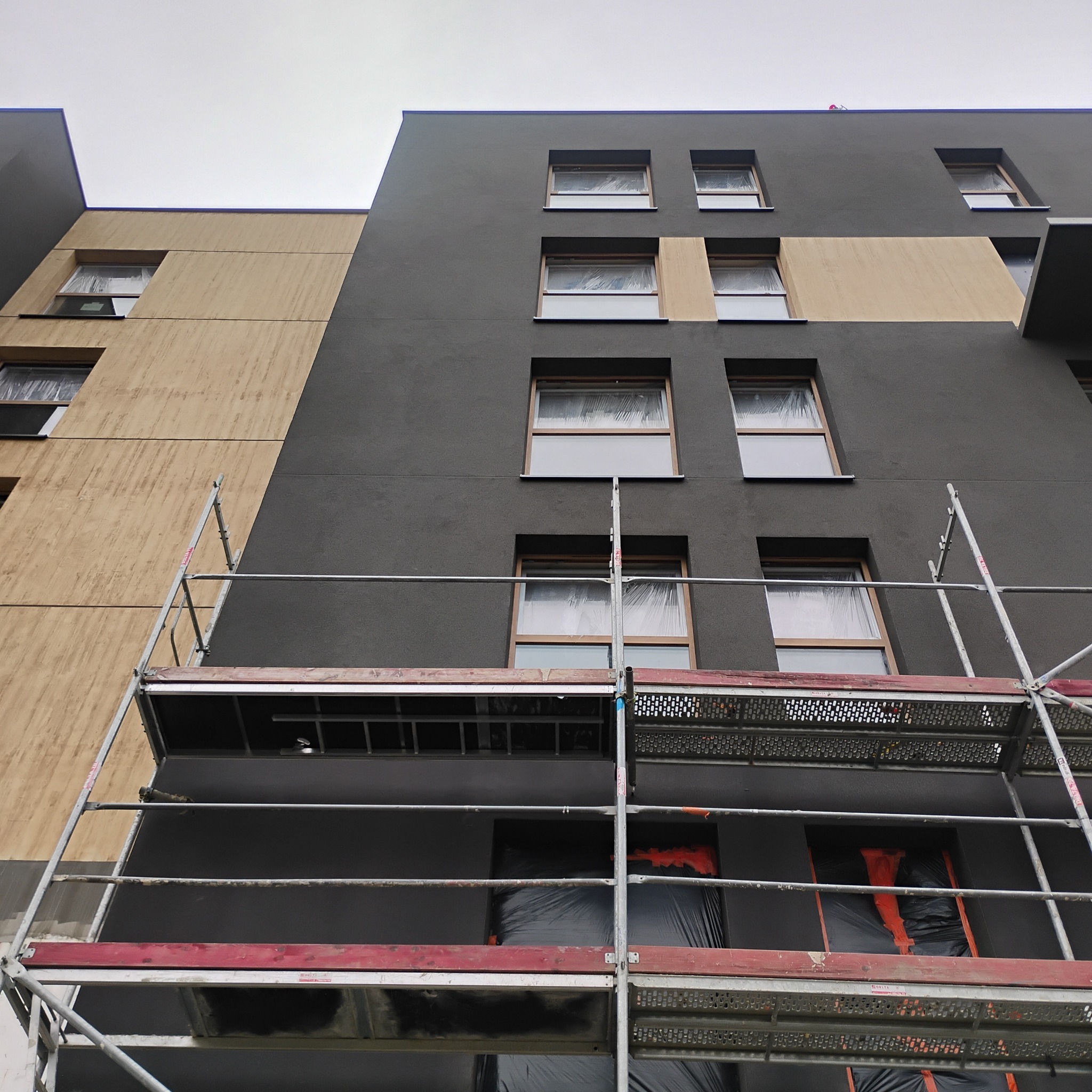 Elewacja budynku w trakcie malowania na szaro i beżowo, okna zabezpieczone folią, widoczne rusztowania. Ujęcie z dołu.