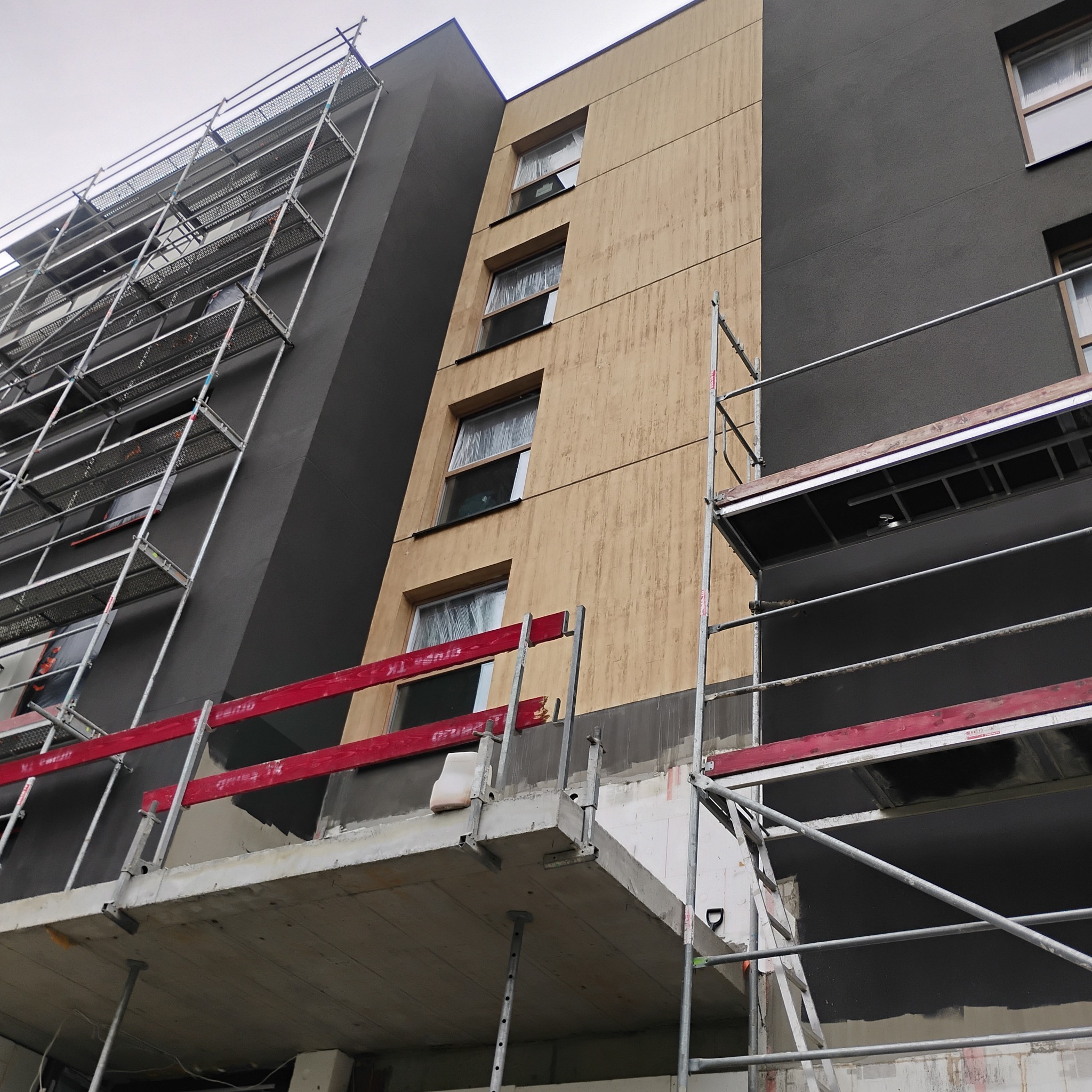 Ocieplanie i malowanie elewacji budynku wielorodzinnego z użyciem rusztowań. Widoczne fragmenty ocieplonej i pomalowanej elewacji w odcieniach beżu i szarości.