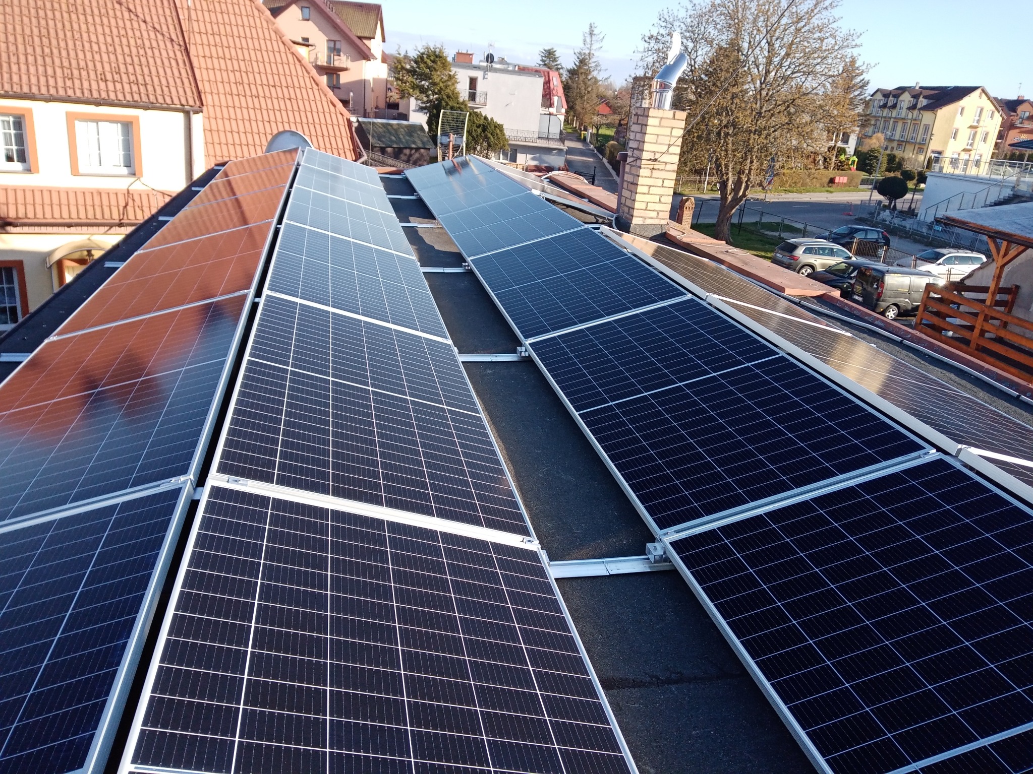 Instalacja fotowoltaiczna na dachu domu jednorodzinnego, widok z góry, panele ułożone rzędami, w tle zabudowa mieszkalna i błękitne niebo.