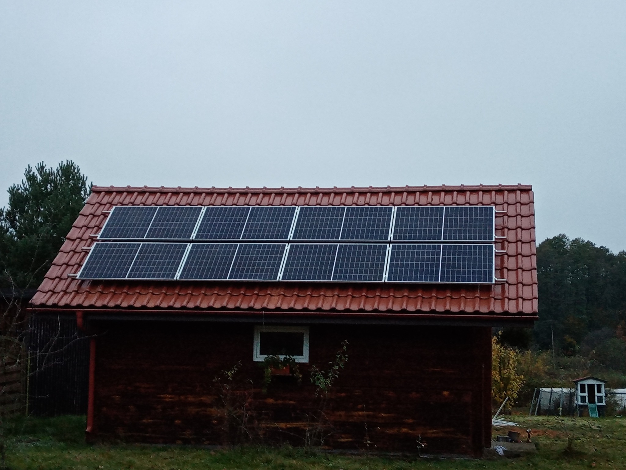 Czerwony dach z panelami słonecznymi na drewnianym domku w jesiennej scenerii, widok z dołu, pochmurne niebo.