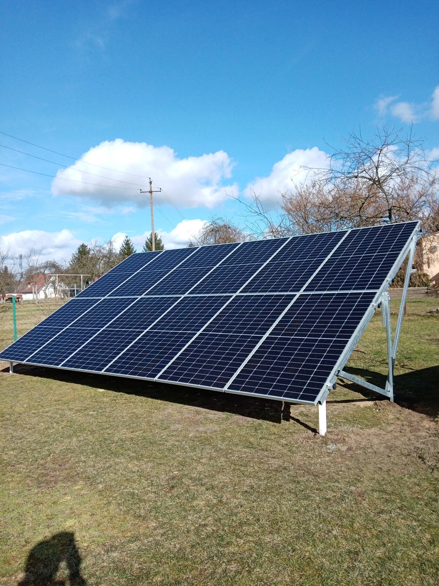 Panel fotowoltaiczny na trawie, wsparty na metalowej konstrukcji, błękitne niebo z chmurami w tle. Widoczna cień osoby robiącej zdjęcie.