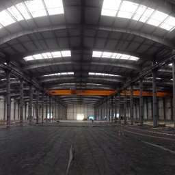 AERO MICHAŁ SUCHANEK - Wnętrze hali produkcyjnej w trakcie budowy. Widoczne stalowe konstrukcje, świetliki w dachu i czarna folia na podłożu. W tle suwnica.