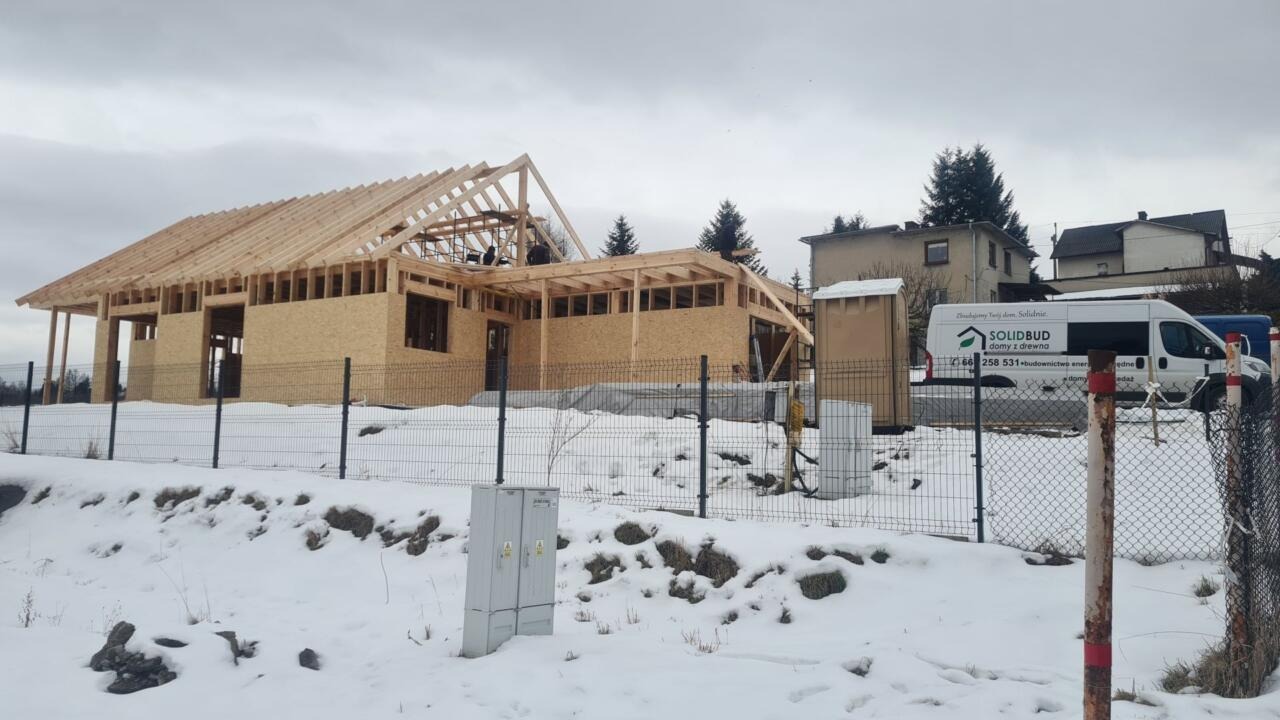 Budowa domu szkieletowego w zimowej scenerii. Widoczna konstrukcja ścian z płyt OSB i drewniany szkielet dachu. Na drugim planie budynek i samochód firmy budowlanej.