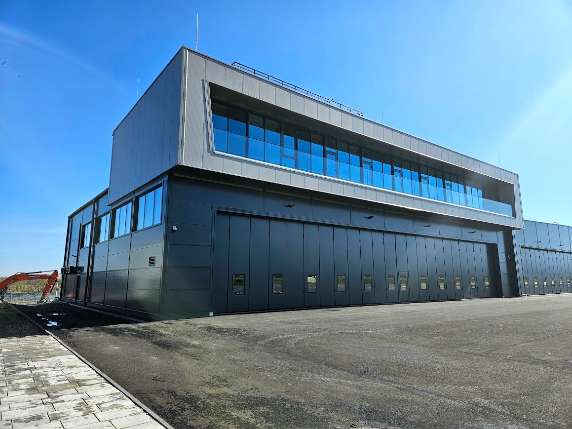 Nowoczesny, dwukondygnacyjny budynek przemysłowy z szarymi panelami i dużymi oknami, z pomarańczową koparką w tle, na tle błękitnego nieba.
