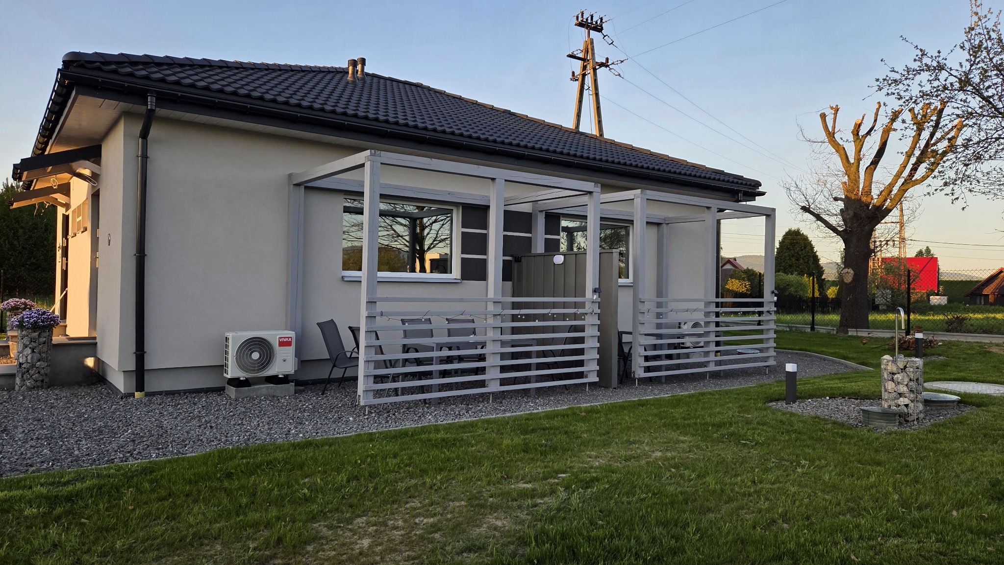 Nowoczesny, parterowy dom z szarą elewacją, drewnianą pergolą i klimatyzatorem. Widok na zadbany trawnik i elementy dekoracyjne z kamienia.