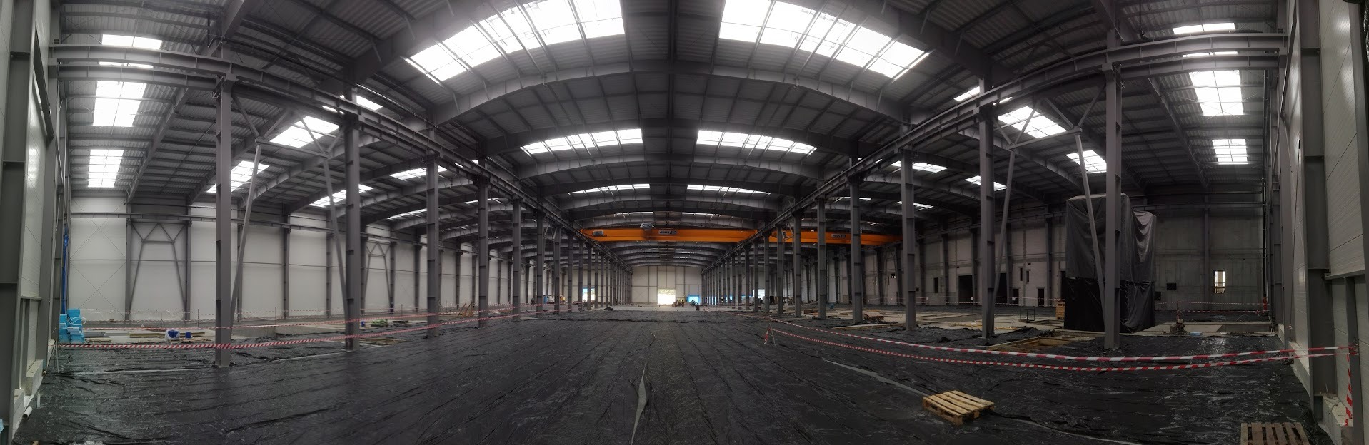 Wnętrze hali produkcyjnej w trakcie budowy. Widoczne stalowe konstrukcje, świetliki w dachu i czarna folia na podłożu. W tle suwnica.