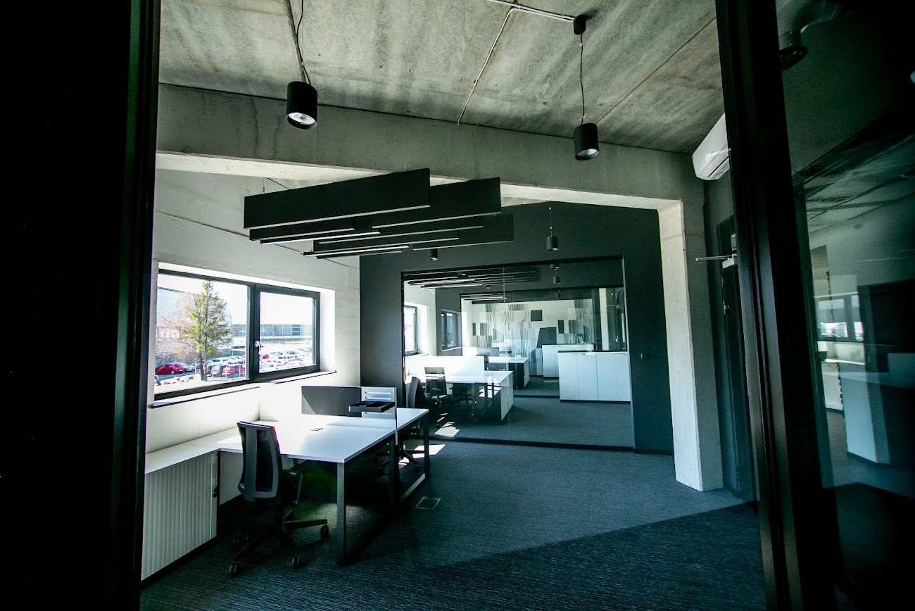 Nowoczesne biuro z surowym betonowym sufitem, czarnymi lampami i białymi biurkami, widoczne przez ciemne obramowanie drzwi. Duże okna wpuszczają naturalne światło.