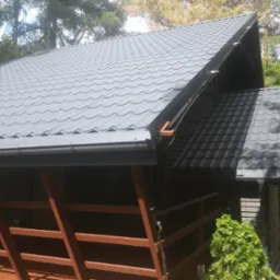 Szary dach z blachy trapezowej na drewnianym domku z brązową balustradą, widoczny fragment orynnowania i otaczająca zieleń. Ujęcie z dołu pod kątem.