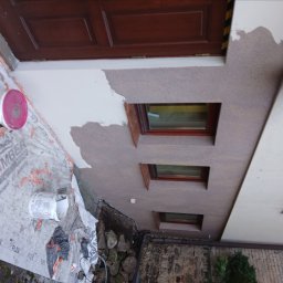 Łuk-Bud - Fragment elewacji z oknami, częściowo pokrytej tynkiem, z widocznymi puszkami farby i zabezpieczeniem podłoża. Prace wykończeniowe w toku.