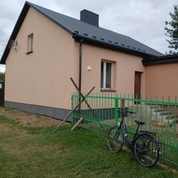 Łuk-Bud - Otynkowany dom jednorodzinny z szarą podmurówką i ciemnym dachem, ogrodzony zielonym płotem. Na trawniku oparty rower i drewniane podpory.