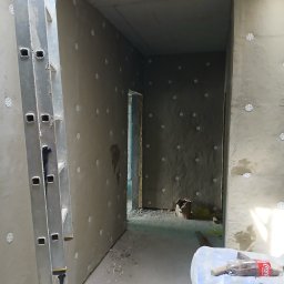 Łuk-Bud - Wnętrze w trakcie ocieplania ścian styropianem, widoczne kołki mocujące, fragment drabiny aluminiowej i worki z zaprawą murarską w pomieszczeniu.