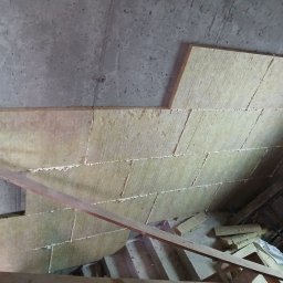 Łuk-Bud - Ocieplenie wełną mineralną pod stropem, widoczne płyty izolacyjne z pianką montażową, fragment drewnianej konstrukcji i surowy beton. Prace izolacyjne w toku.