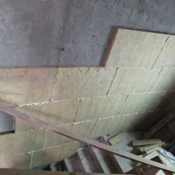 Ocieplenie wełną mineralną pod stropem, widoczne płyty izolacyjne z pianką montażową, fragment drewnianej konstrukcji i surowy beton. Prace izolacyjne w toku.