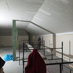 Łuk-Bud - Poddasze w trakcie prac wykończeniowych: widoczne rusztowania, płyty gipsowe na skosach dachu i ścianach, oraz pracownik w oddali.