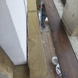 Łuk-Bud - Fragment ocieplonej ściany budynku w trakcie prac, widok z rusztowania. Na ścianie widoczne płyty izolacyjne i warstwa tynku. Częściowo widoczna osoba w kombinezonie roboczym.