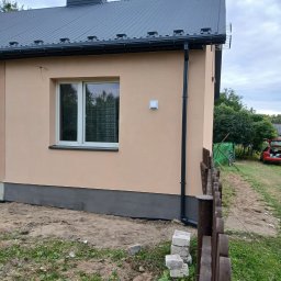 Łuk-Bud - Wykończona elewacja domu jednorodzinnego z beżowym tynkiem, szarą podmurówką i grafitowym dachem. Widoczny fragment drewnianego płotu i ułożone bloczki.