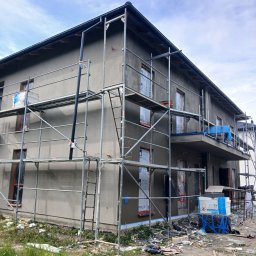 Łuk-Bud - Dom w trakcie tynkowania elewacji, otoczony rusztowaniami. Okna zabezpieczone folią, widoczne materiały budowlane i elementy wykończeniowe. Prace w toku.