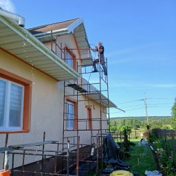 Łuk-Bud - Pracownik myje elewację domu z rusztowania przy użyciu myjki ciśnieniowej. W tle zielony krajobraz i błękitne niebo. Widoczne detale elewacji i narzędzia pracy.