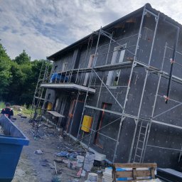 Łuk-Bud - Dom w trakcie ocieplania elewacji, widoczne rusztowania, materiały izolacyjne i pracownik w tle. Etap prac przed nałożeniem tynku.