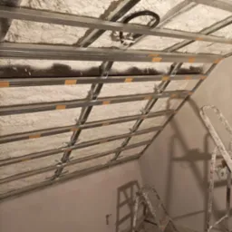 Sufit podwieszany z metalową konstrukcją i pianką izolacyjną, widok z dołu, drabina malarska w rogu. Oświetlenie sufitowe częściowo widoczne.