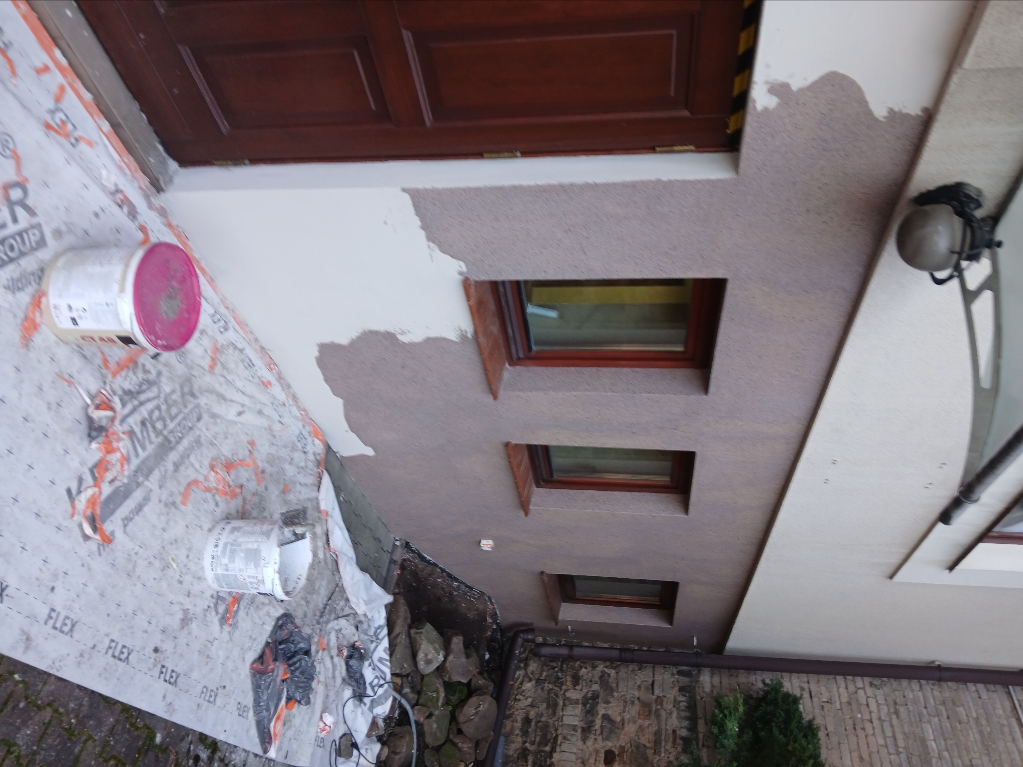 Fragment elewacji z oknami, częściowo pokrytej tynkiem, z widocznymi puszkami farby i zabezpieczeniem podłoża. Prace wykończeniowe w toku.