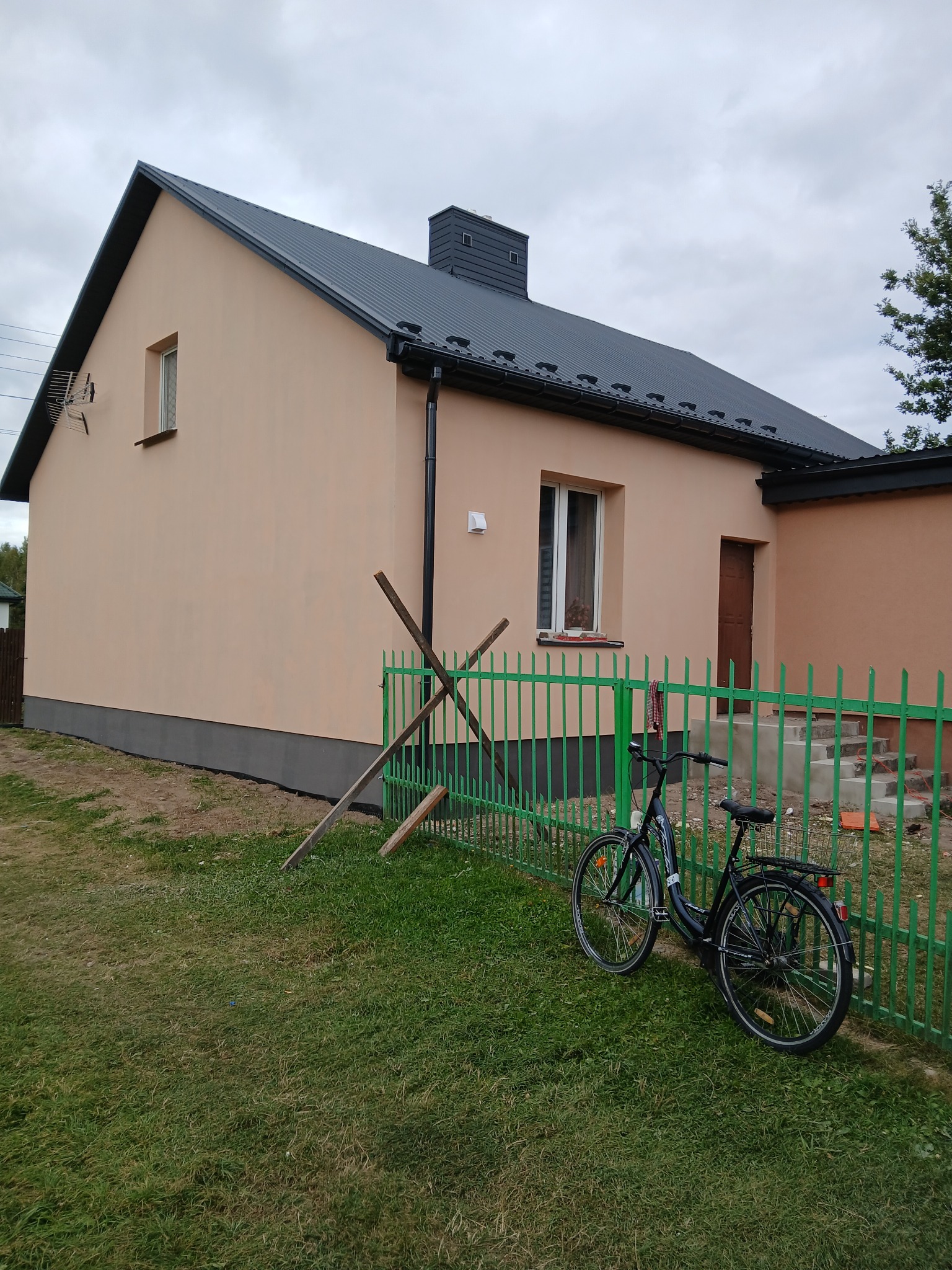 Otynkowany dom jednorodzinny z szarą podmurówką i ciemnym dachem, ogrodzony zielonym płotem. Na trawniku oparty rower i drewniane podpory.