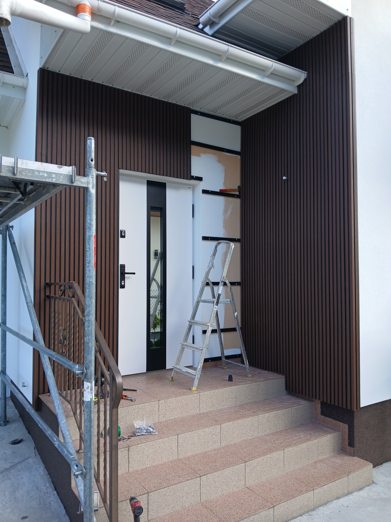 Montaż elewacji z paneli drewnopodobnych przy wejściu do domu. Widoczne rusztowanie, drabina i narzędzia. Częściowo zamontowane panele w kolorze ciemnego brązu.